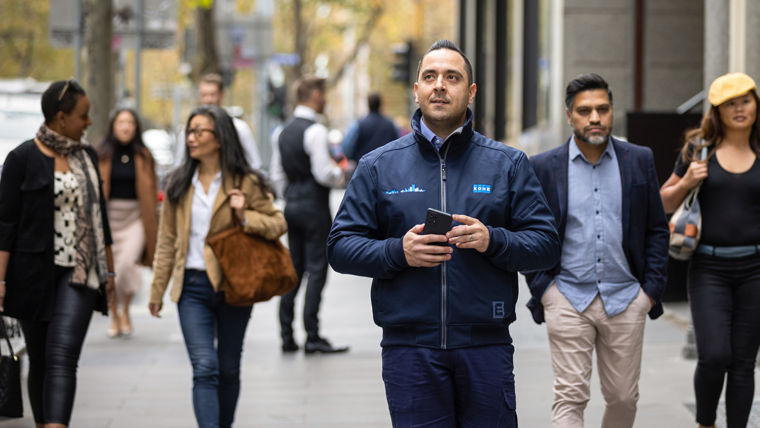 A KONE technician holds a mobile device while standing on a busy urban street.